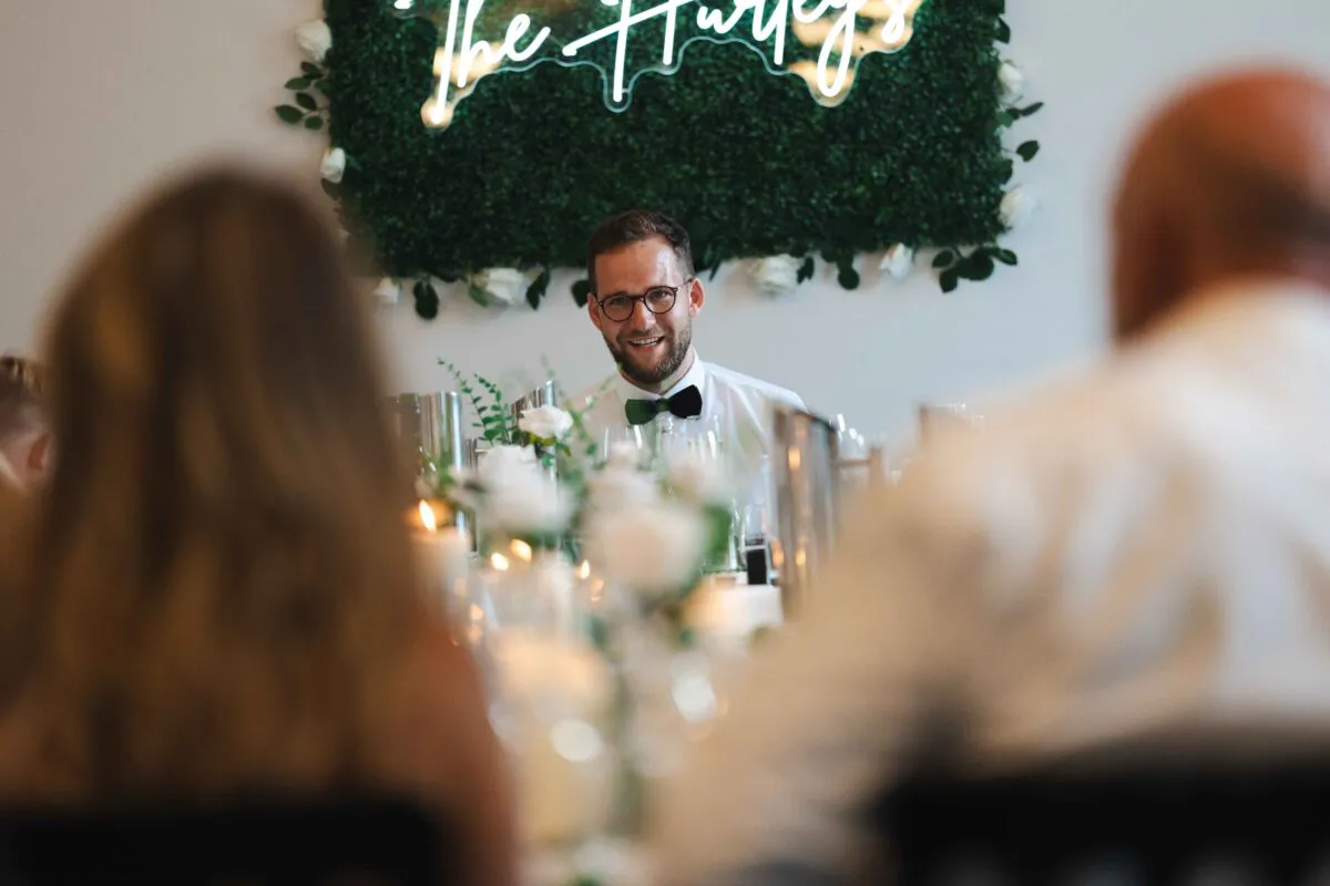 A smiling groom seated at the top table during wedding speeches, framed by guests in the foreground and a neon sign against a greenery wall.