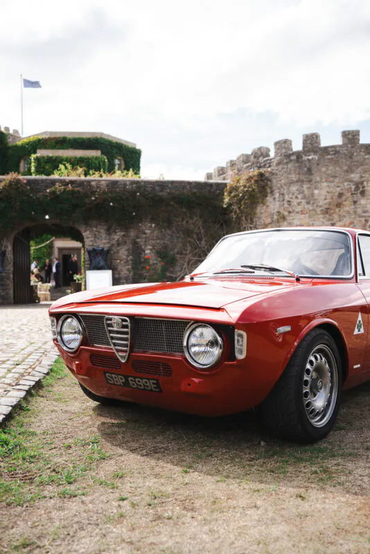 A vintage red Alfa Romeo classic wedding car is parked neatly on a cobbled courtyard outside an ancient stone castle venue.