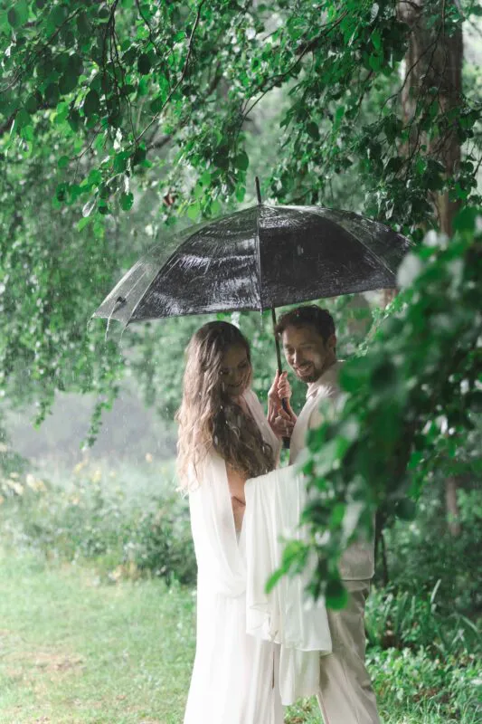 A newlywed couple embraces lovingly underneath a black umbrella while standing in a lush, rainy woodland surrounded by vibrant green foliage.