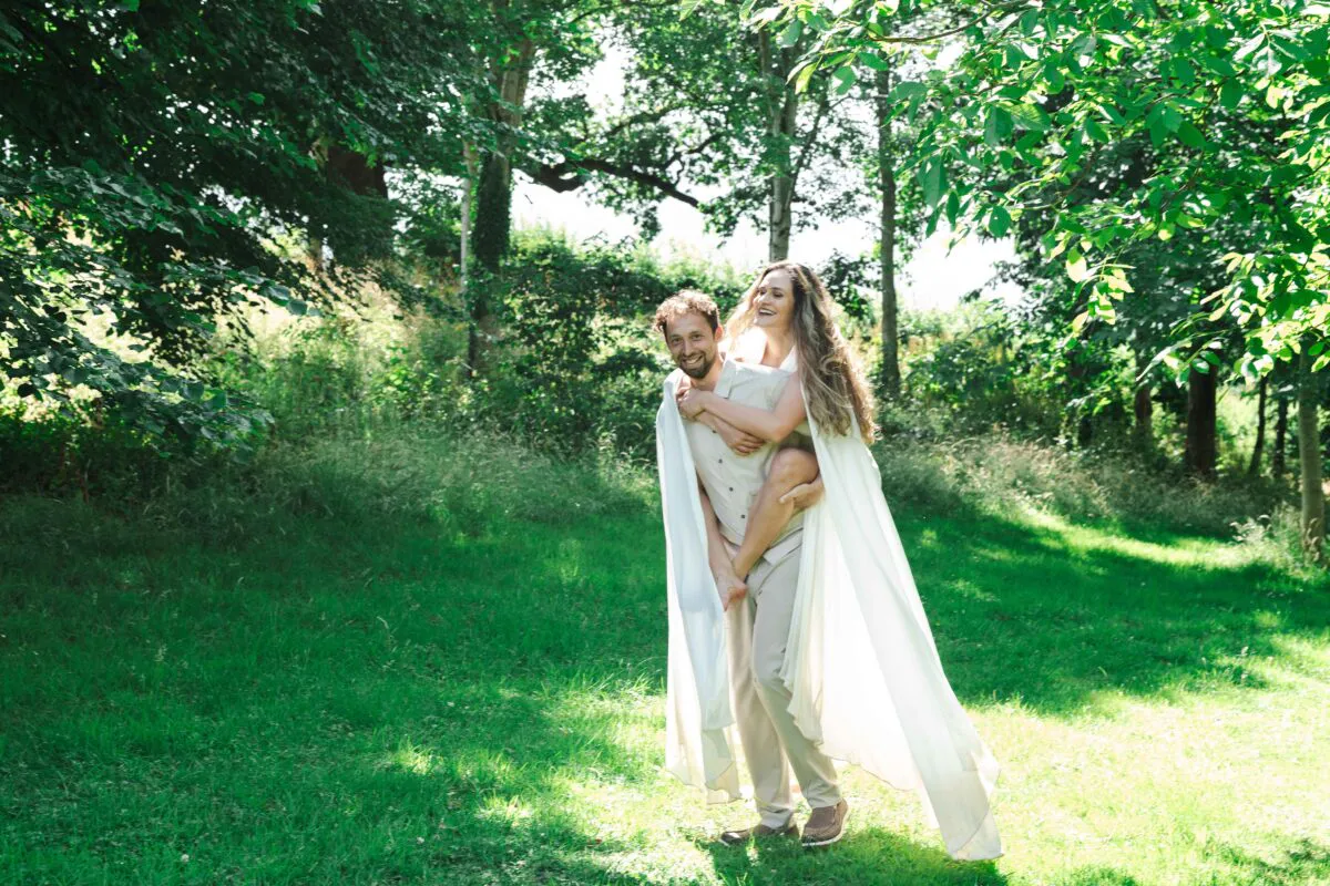 A groom gives his bride a playful piggyback ride through a sun-dappled woodland while laughing together under a bright summer sky.