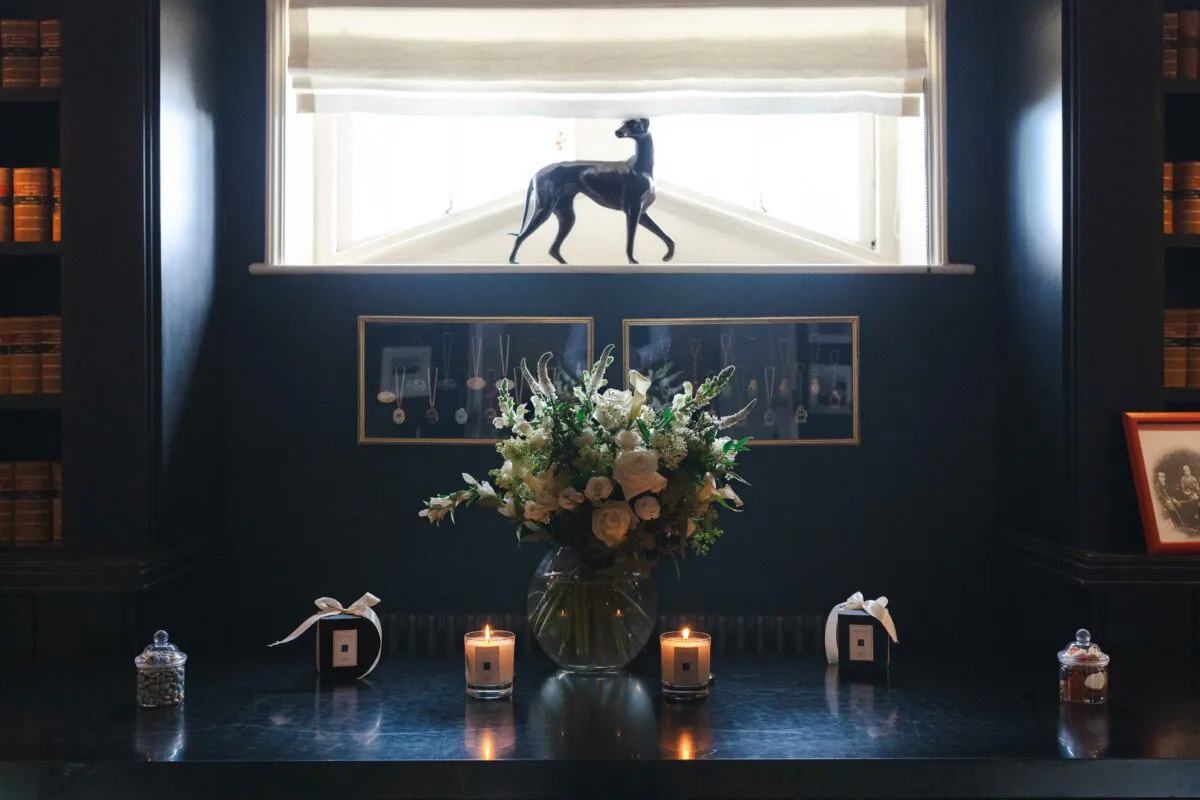 An atmospheric library interior features a dark wooden table styled with a lush white floral arrangement in a glass vase, flanked by lit candles and gift boxes under a window.