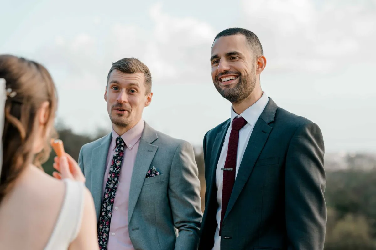 Two smiling groomsmen in tailored suits share a light-hearted, joyful moment while interacting with a guest during the wedding reception.