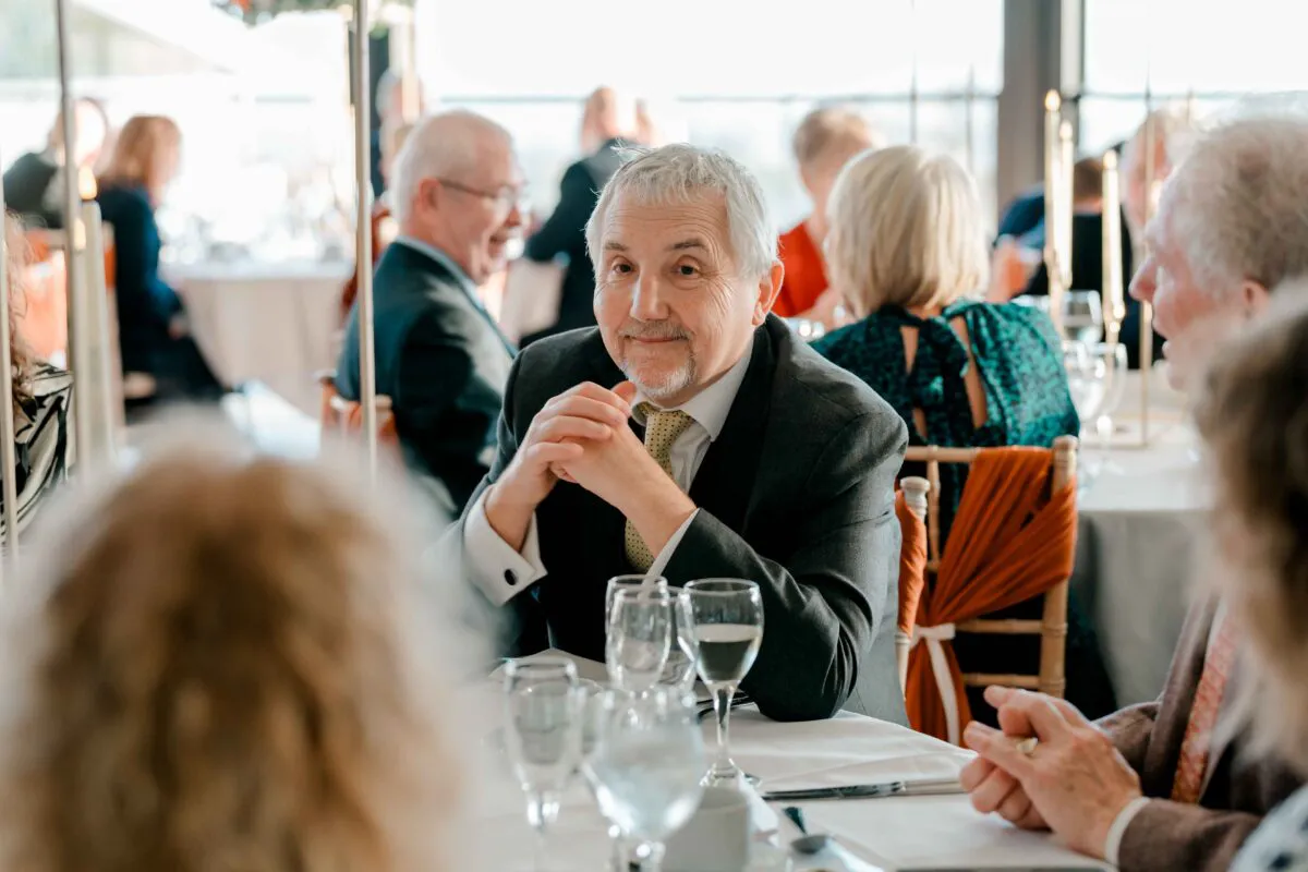A mature male guest wearing a grey suit and patterned tie sits at a wedding breakfast table, smiling towards the camera with his hands clasped.