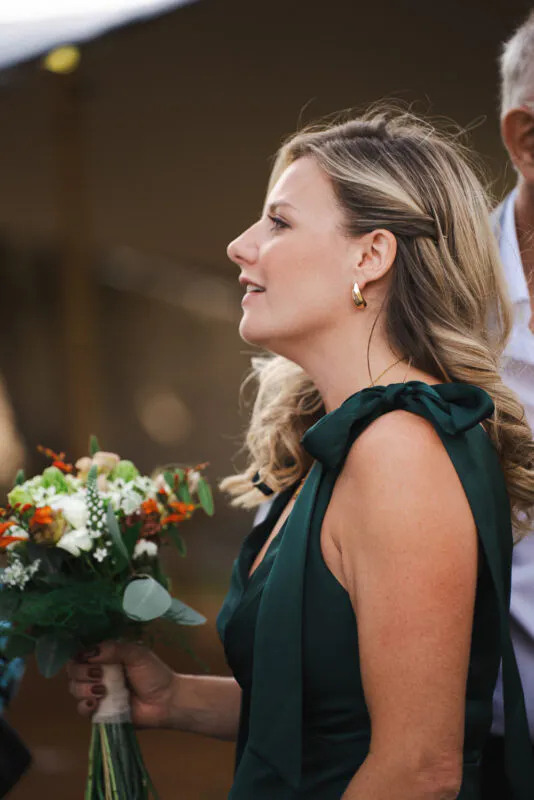 A blonde bridesmaid in a dark green dress with bow-detailed shoulder straps holds a hand-tied bouquet during an outdoor wedding ceremony.