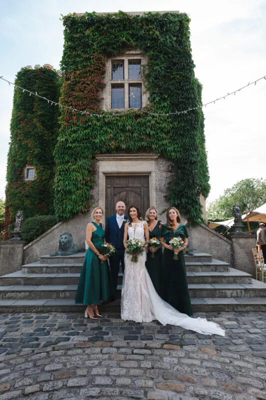The bride and groom pose on stone steps with four bridesmaids wearing emerald green dresses in front of an ivy-covered stone tower.
