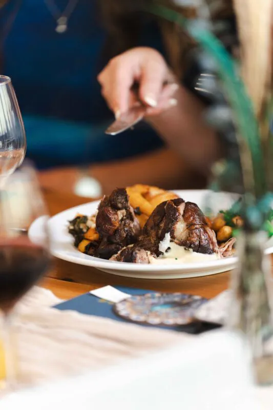 A guest at a wedding reception cuts into a beautifully plated meat dish with roasted potatoes during the wedding breakfast.