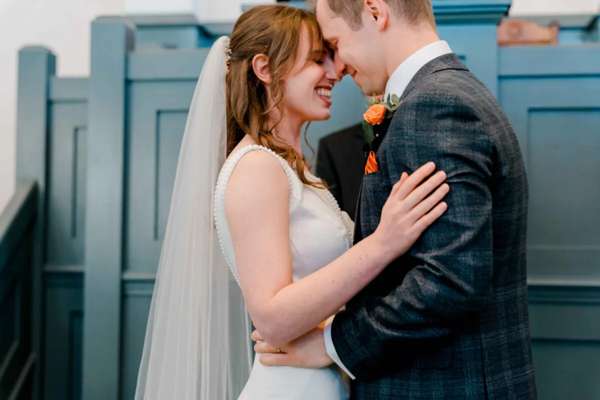 A newlywed couple share a joyful nose-to-nose moment indoors against a blue panelled backdrop, with the bride wearing a pearl-detailed dress and veil.