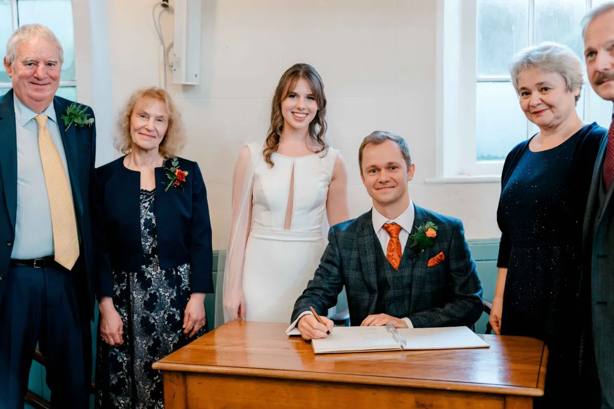 The groom signs the wedding register seated at a wooden desk, flanked by the smiling bride, parents, and grandparents during the ceremony.