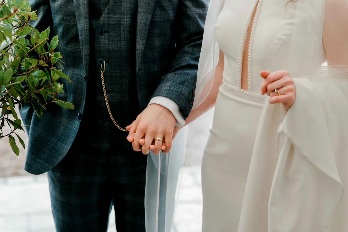 A bride and groom hold hands tightly, showcasing their wedding rings and a classic pocket watch chain attached to the groom's tweed waistcoat.