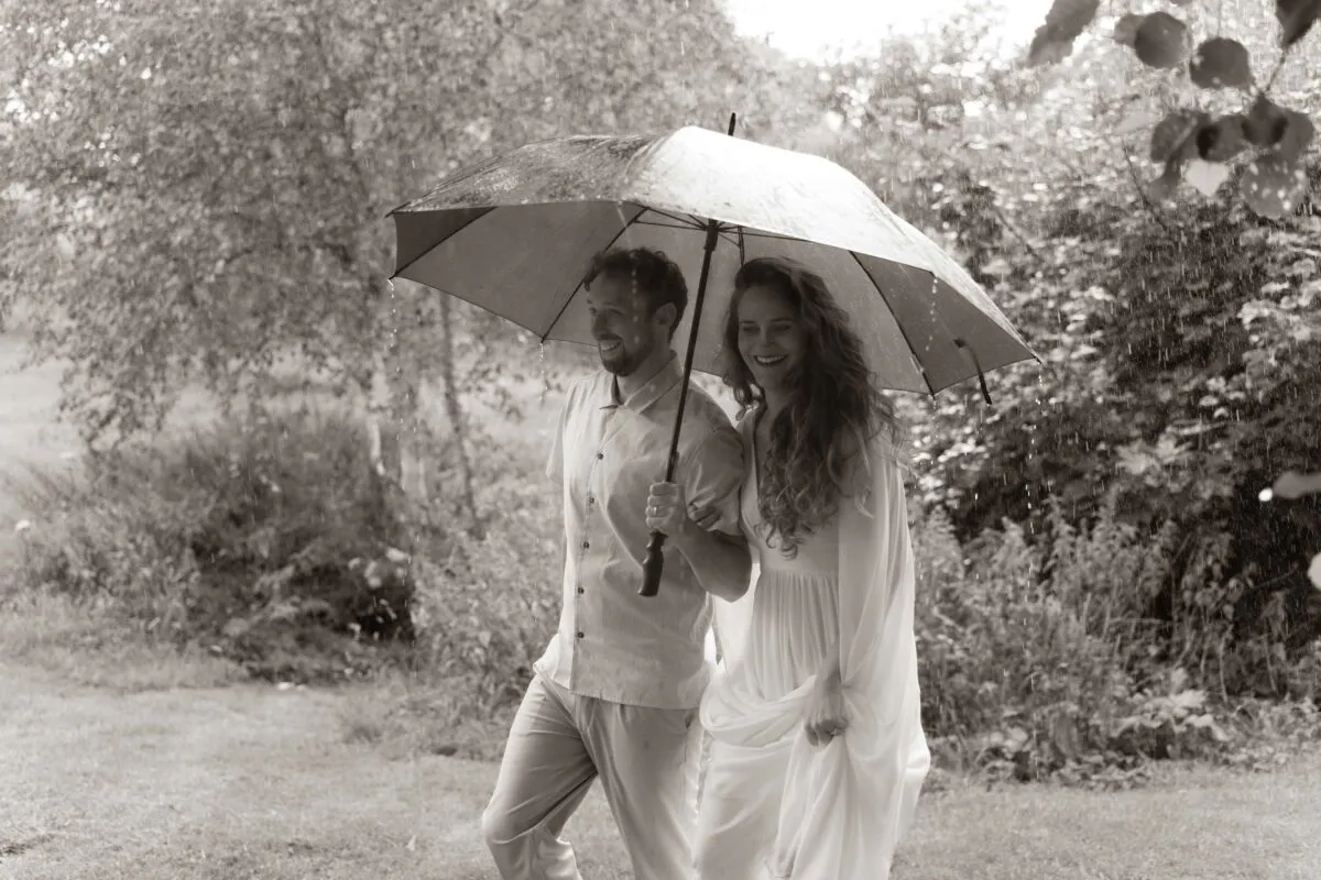 A bride and groom share a joyful moment walking together under an umbrella during a rain shower in a woodland setting.