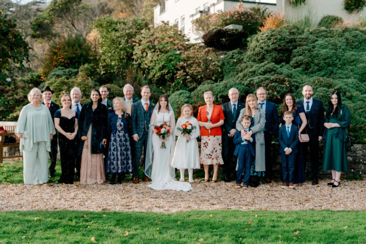 A large wedding party and family members pose for a formal group portrait on a gravel path outdoors during a gentle rain shower.