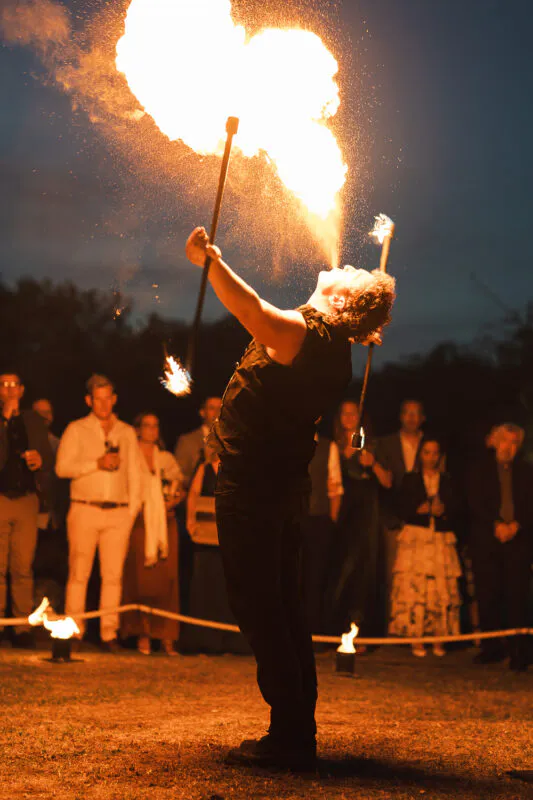 A fire performer creates a spectacular plume of fire against the night sky while wedding guests watch in awe.