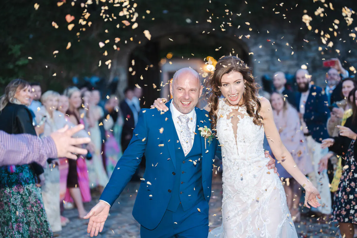 A joyful newlywed couple walks through a cheering crowd of guests who are showering them with confetti in front of an ancient stone archway.