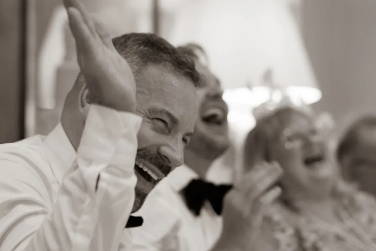 A guest in a white shirt and black bow tie laughs heartily and claps his hands during the wedding speeches.