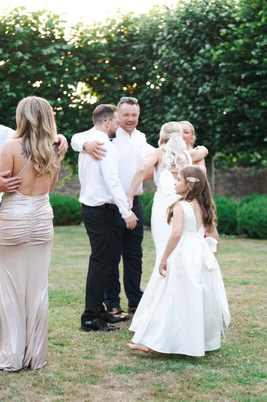 Guests and family members mingle and embrace outdoors on a grassy lawn during a relaxed wedding reception in the warm afternoon light.