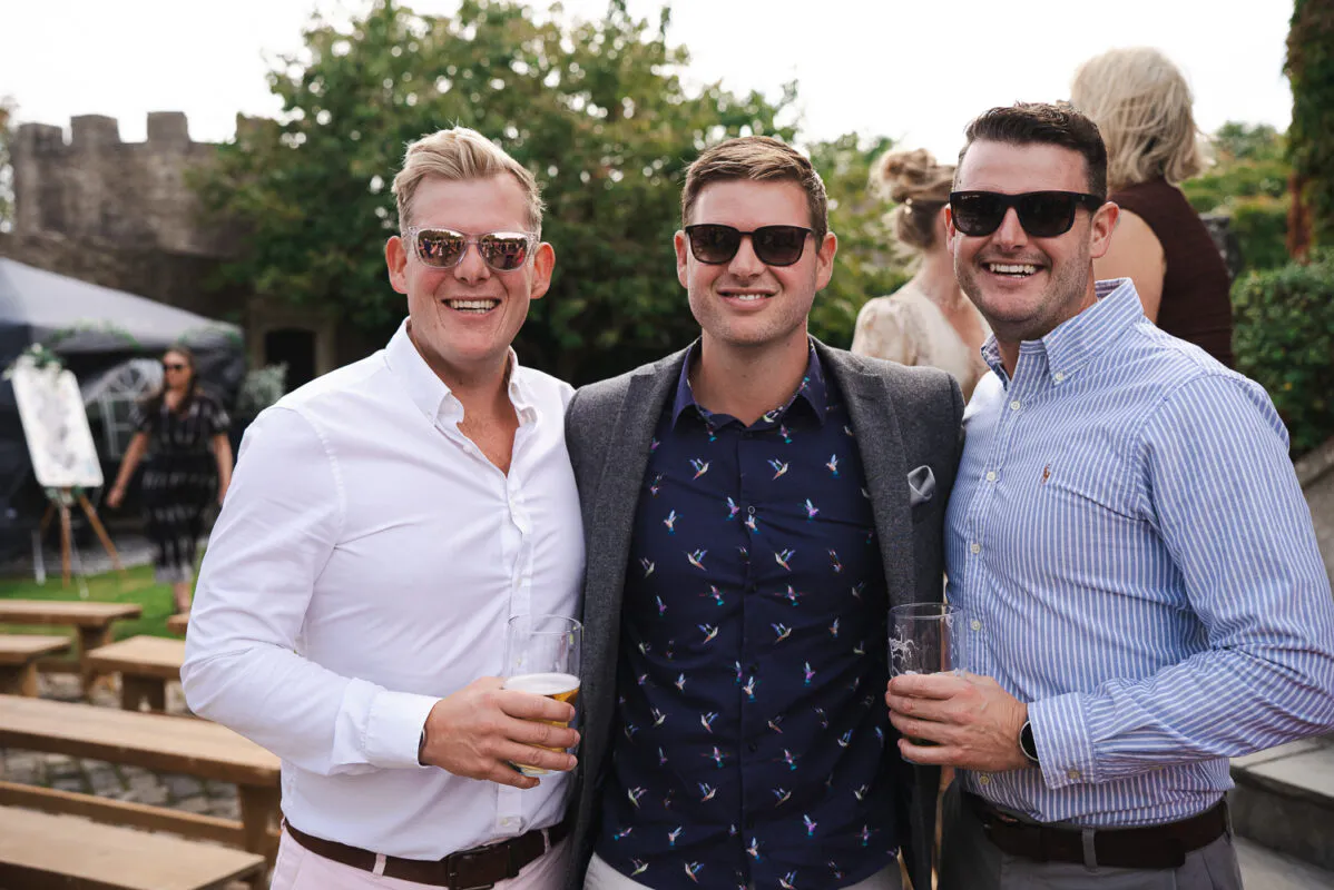 Three smiling male wedding guests wearing sunglasses and smart-casual shirts stand together outdoors holding drinks during a sunny wedding reception.
