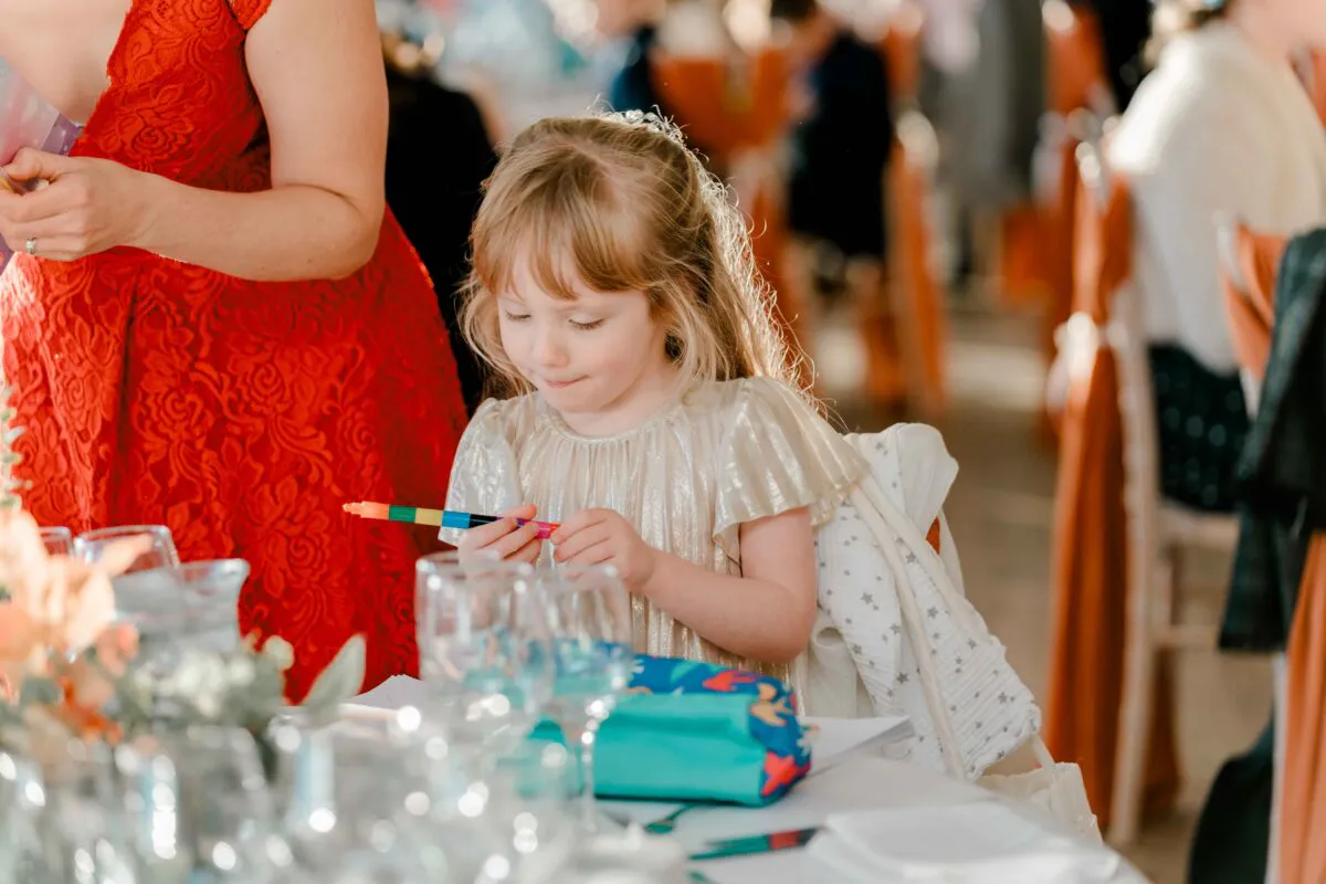 A young flower girl sits attentively at a wedding reception table, holding coloured pens while seated next to a guest in a vibrant red dress.