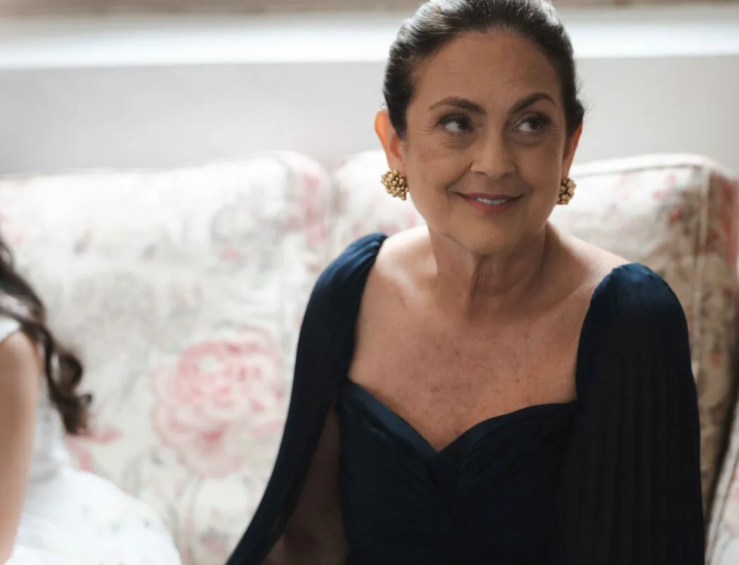 An elegant mother of the bride smiling warmly while seated on a floral patterned sofa, wearing a dark blue gown and gold cluster earrings.
