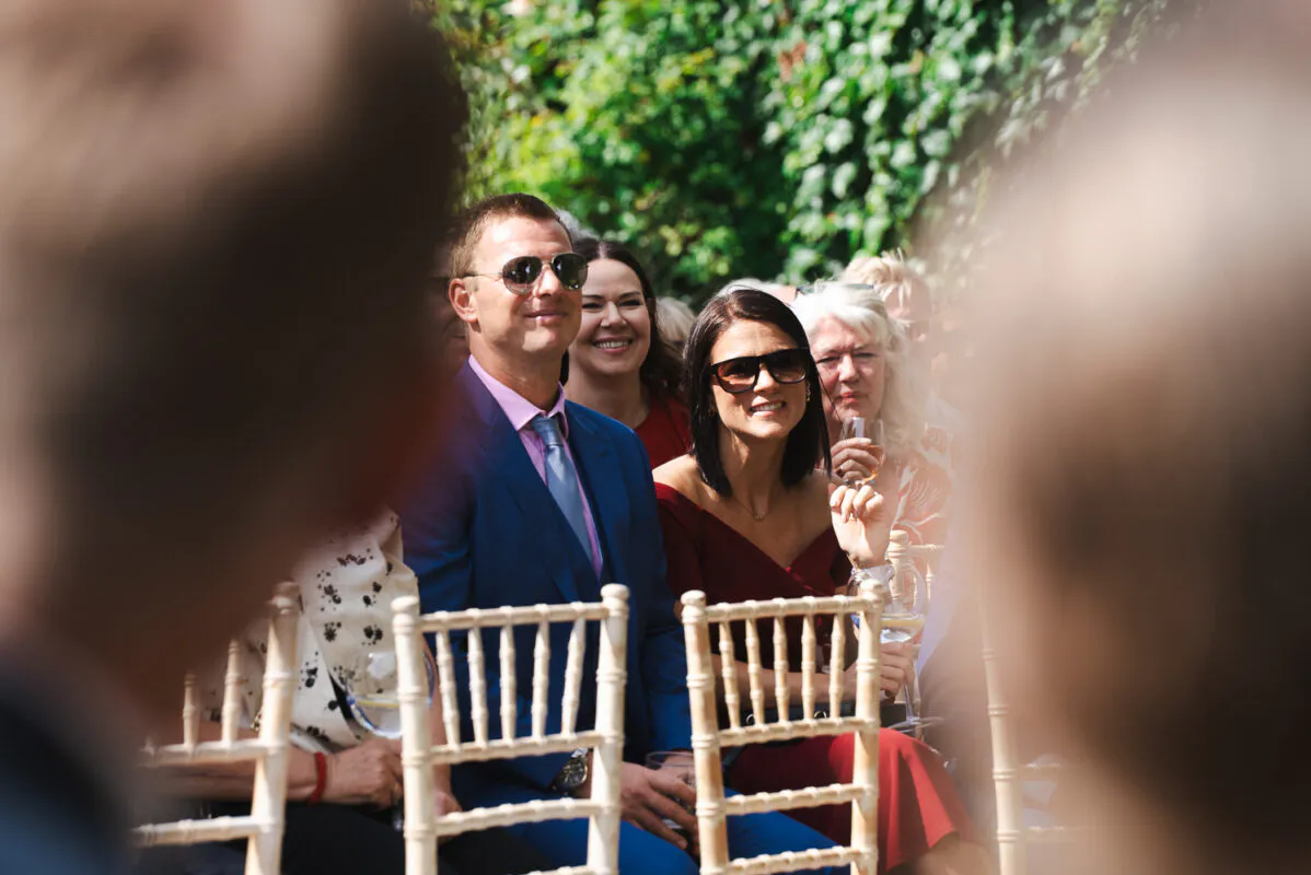 A candid shot of wedding guests seated outdoors on Chiavari chairs, enjoying the sunshine with drinks in hand while wearing sunglasses.