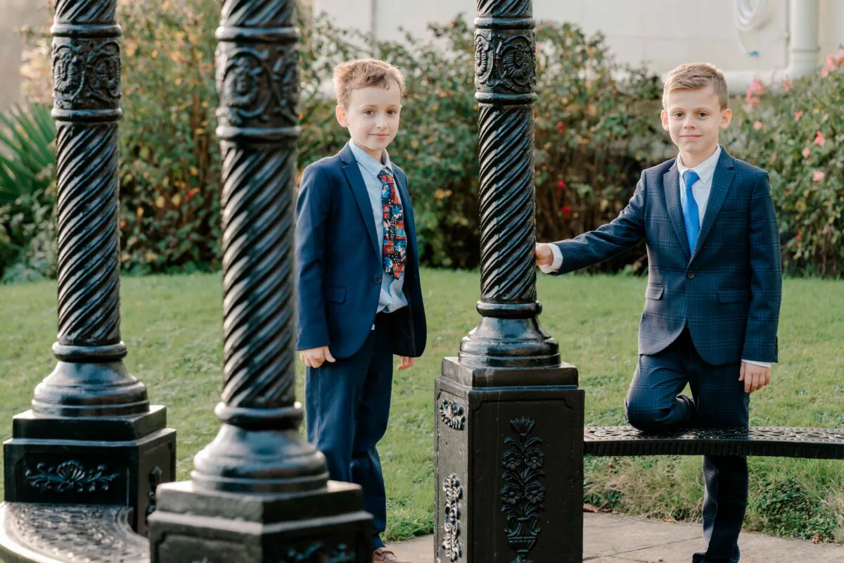 Two page boys in smart navy suits pose near ornate black cast iron pillars in a lush garden setting.