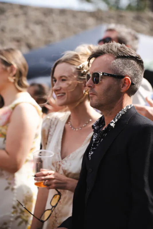 Smiling guests enjoy a sunny outdoor wedding reception, with a man wearing sunglasses and a patterned shirt standing next to a laughing woman holding a drink.