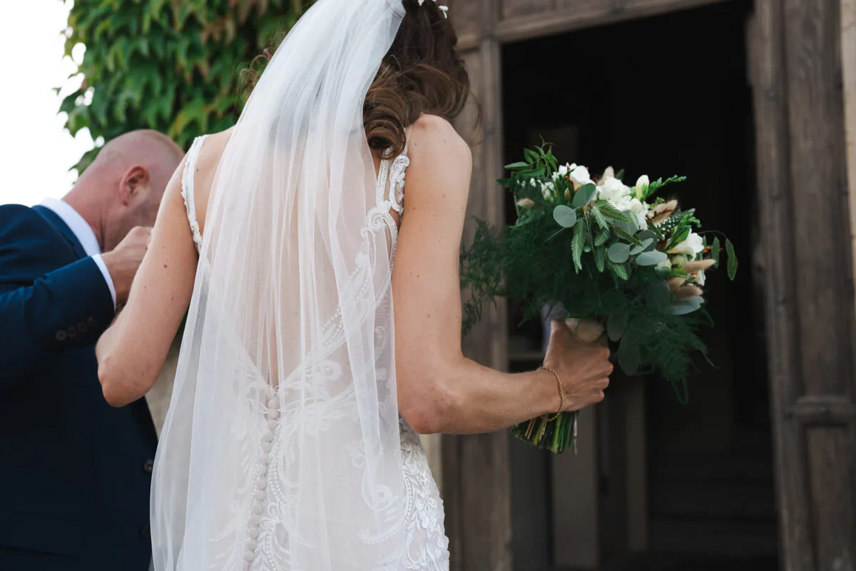 A groom gently kisses the hand of his bride as they walk away together outside a rustic stone church doorway.