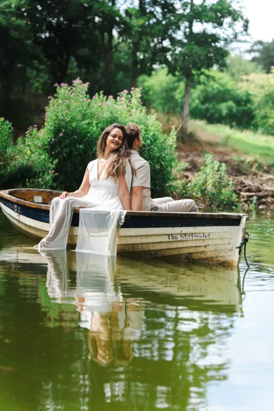 A bride and groom sit back to back in a wooden rowing boat named The Shrimplet on a calm lake, surrounded by lush green foliage.