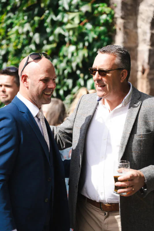 Two male wedding guests in smart suits laugh and chat outdoors during the drinks reception in front of an ivy-covered wall.