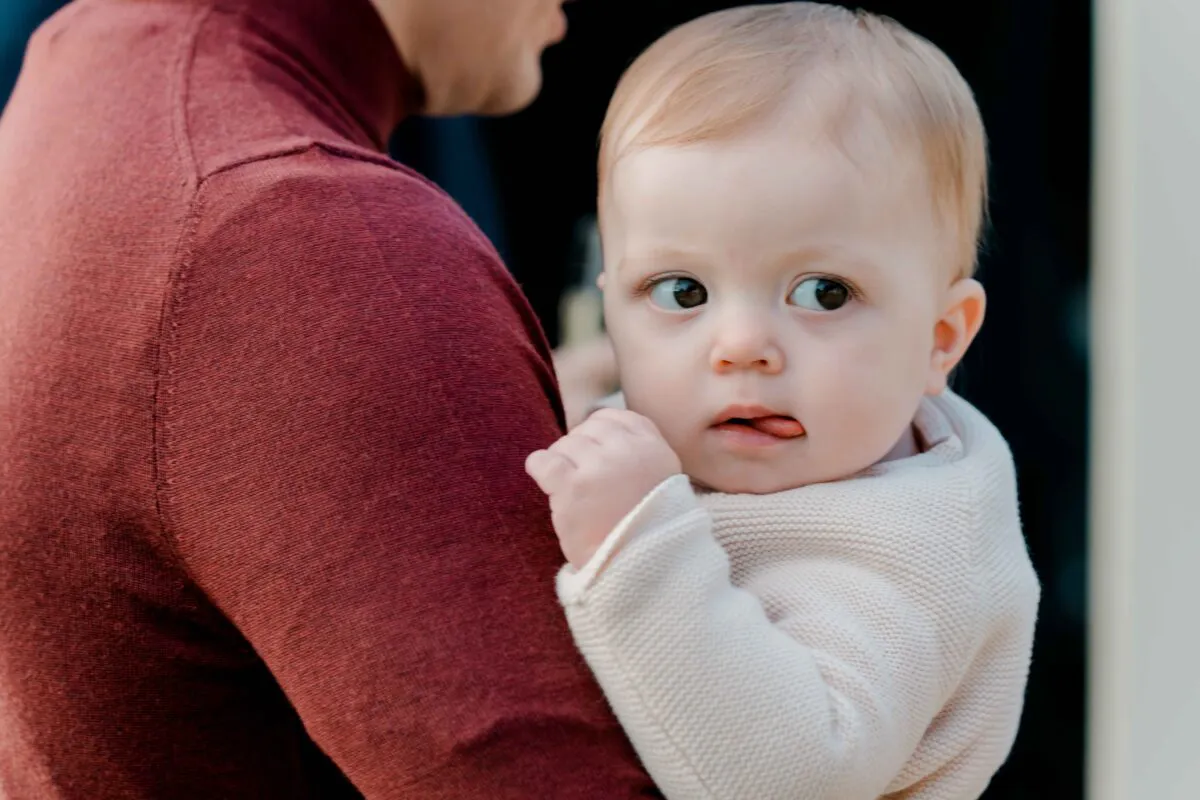 An adorable baby with light hair and a cream jumper is held in the arms of a guest wearing a maroon turtleneck sweater.