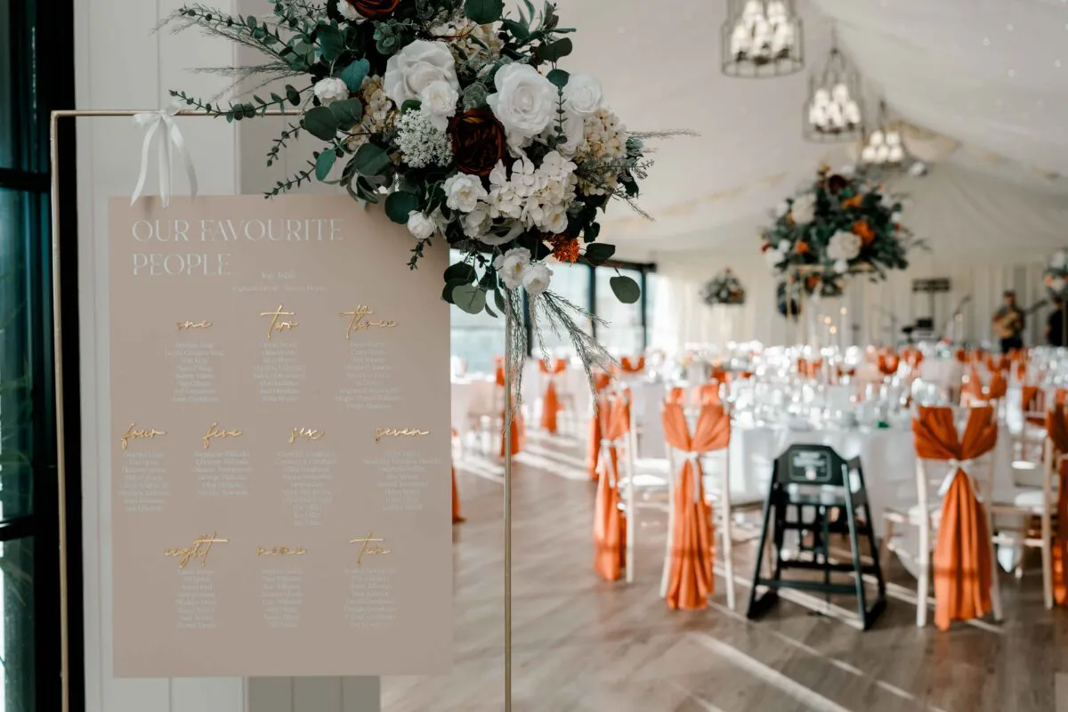 An elegant wedding seating plan sign featuring gold calligraphy and a rich floral arrangement stands in the foreground of a brightly lit marquee reception venue with orange-draped chairs.