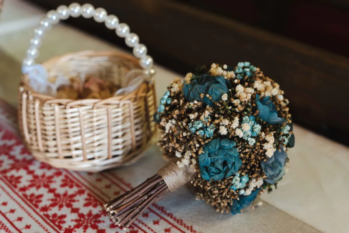 A rustic dried flower bouquet featuring blue and neutral tones sits beside a woven basket with a pearl handle on a patterned table runner.