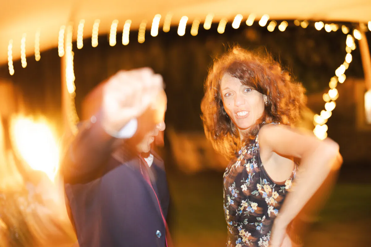 A joyful woman and man energetically dance at a lively evening wedding reception illuminated by overhead string lights.