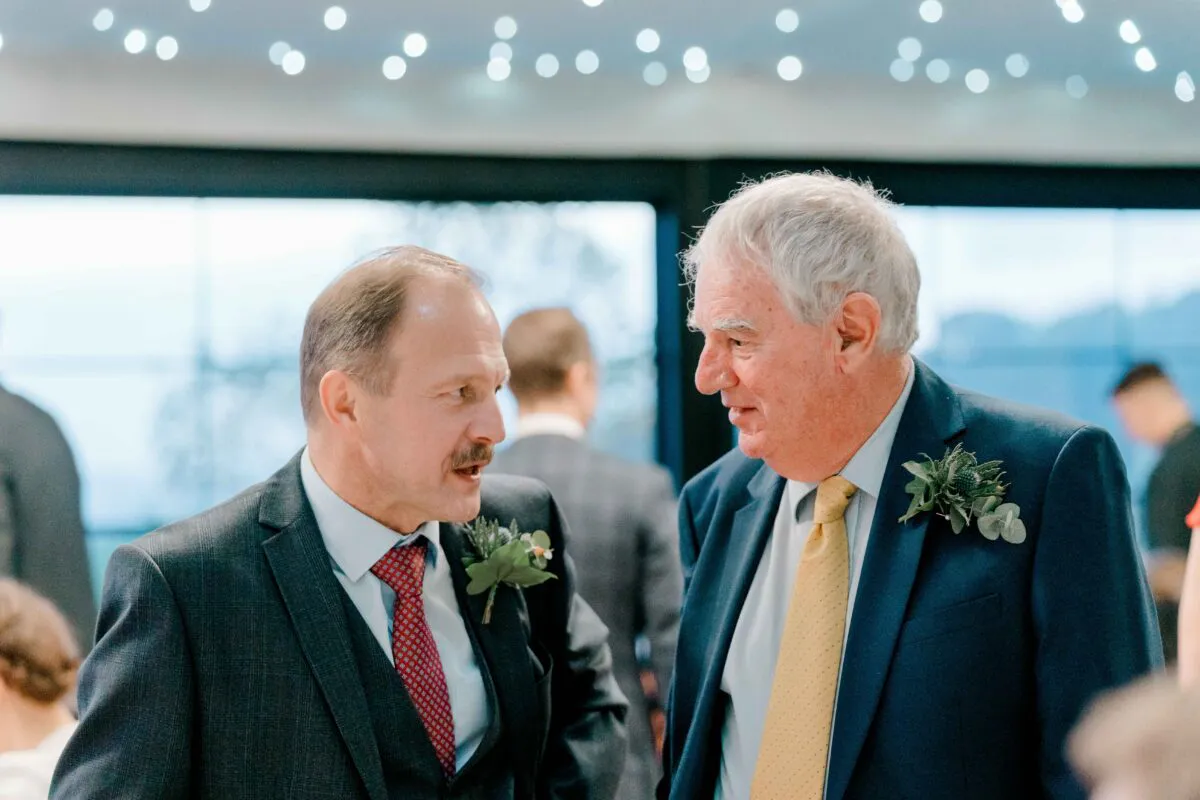 Two older wedding guests in tailored suits converse happily together during the reception beneath a twinkling fairy light canopy.