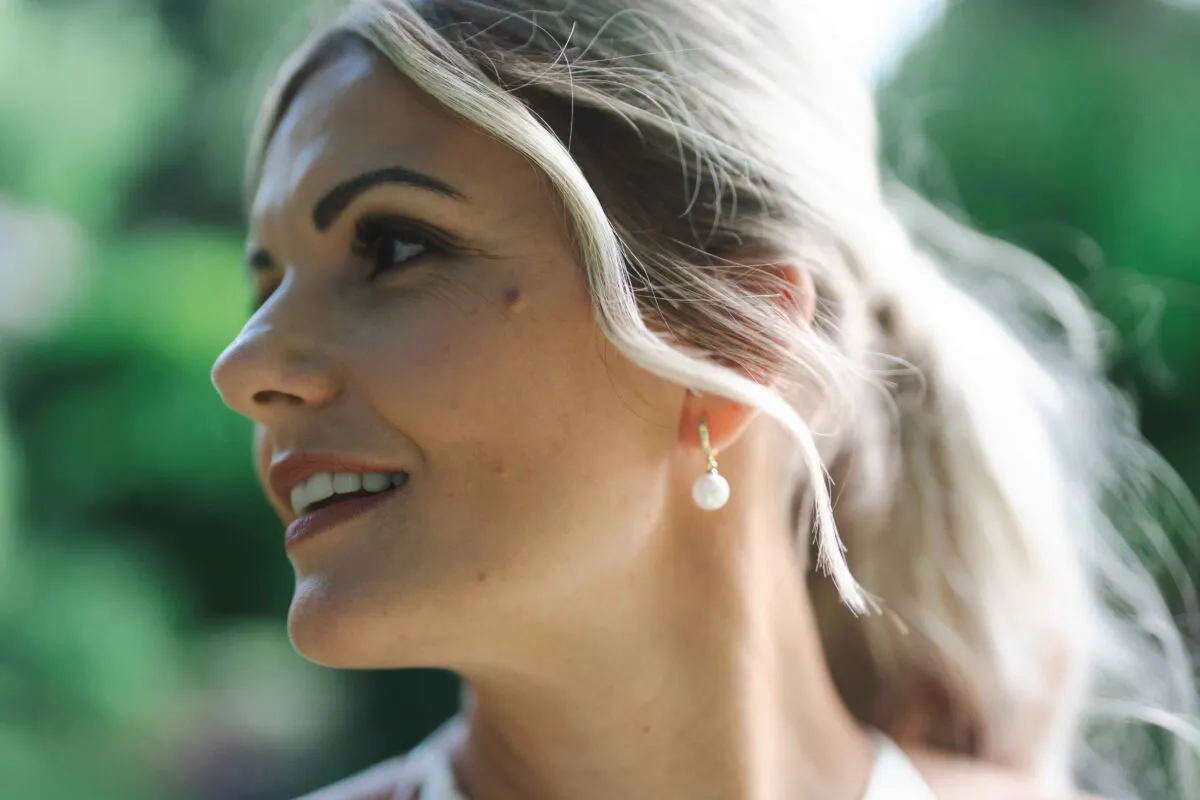 A close-up portrait of a blonde bride with a low ponytail and pearl drop earrings looking away with a soft smile outdoors.