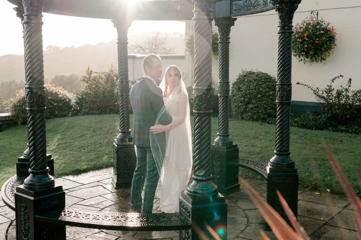 A newlywed couple stands inside an ornate black metal bandstand during golden hour, framed by decorative columns and a soft sun flare.
