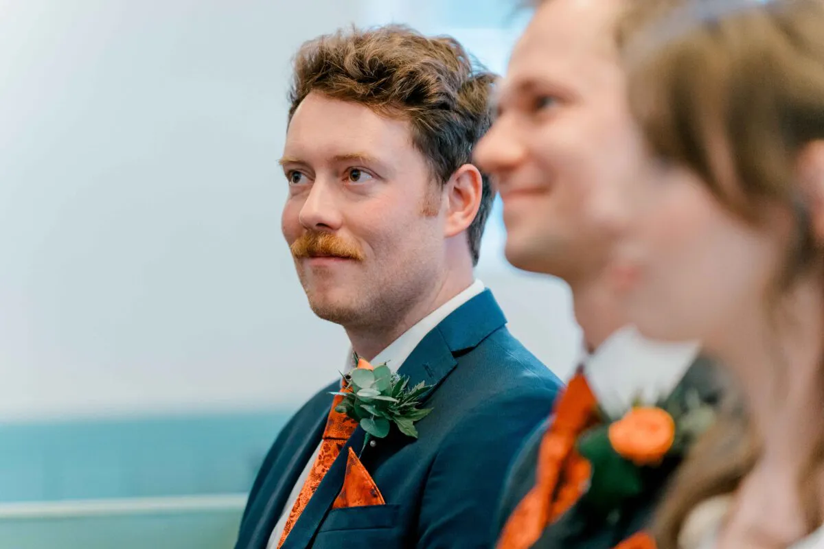 A groom with a ginger moustache and a blue suit featuring an orange patterned tie and boutonniere looks on emotionally during the wedding ceremony.