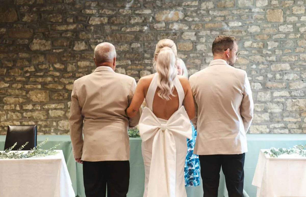 The bride stands with her father and groom from behind, showcasing her halter neck gown with an oversized back bow against an exposed stone wall.
