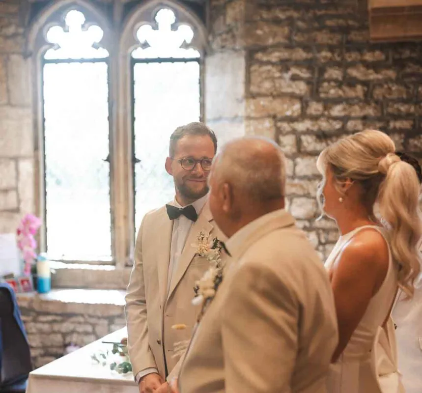 A groom smiling warmly inside a stone church with arched windows during the wedding ceremony, flanked by the father of the bride and the bride.