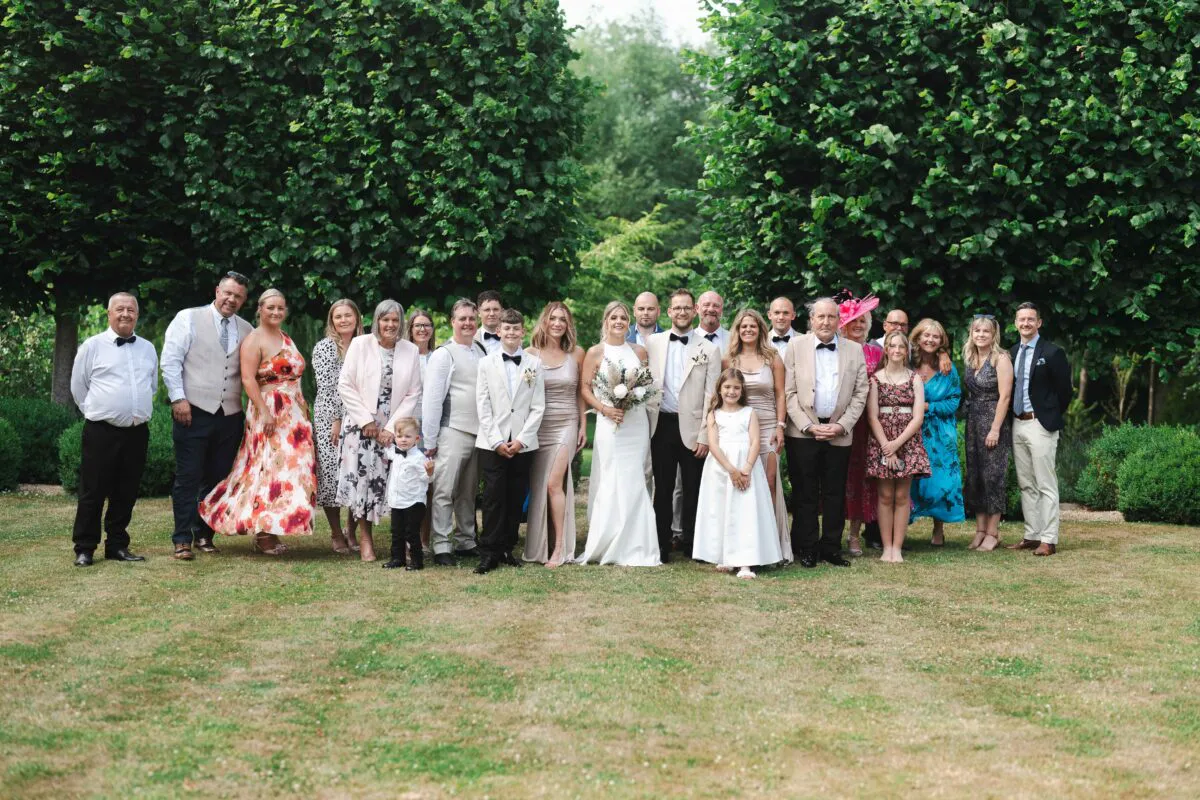 A large wedding party and family group pose together outdoors on a green lawn framed by tall trees during a summer celebration.