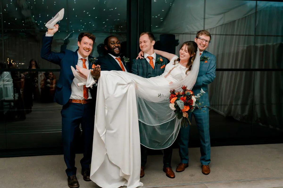 The groom and groomsmen playfully lift the laughing bride horizontal in the air while she holds her cascading orange floral bouquet.