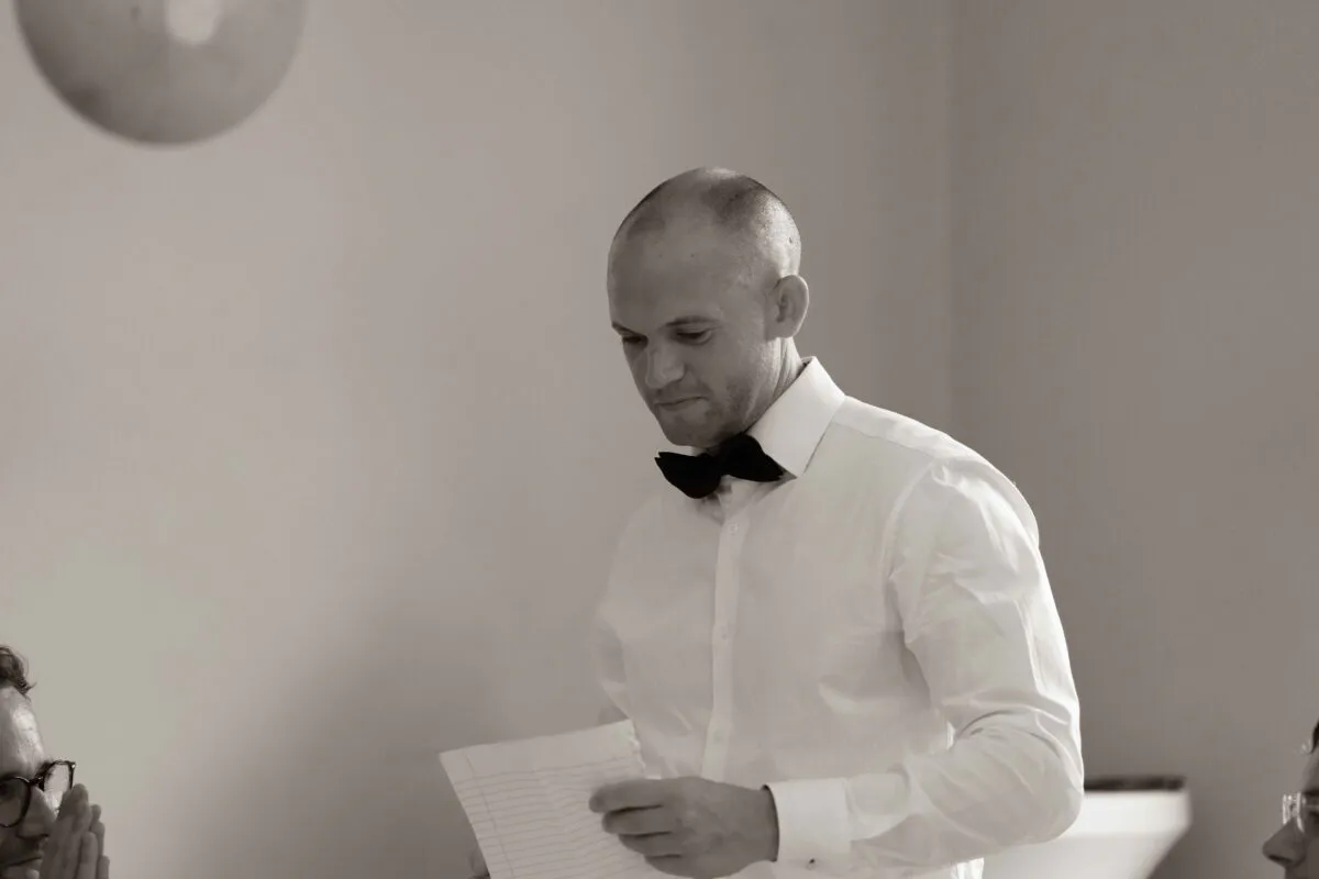 A groom dressed in a white shirt and black bow tie looks down at his speech notes during the wedding reception.