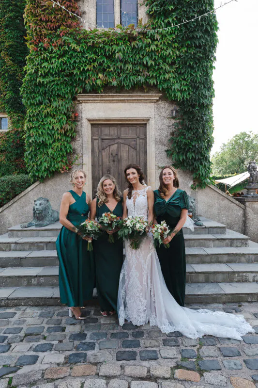 A smiling bride in a lace mermaid wedding dress and her three bridesmaids in rich emerald green dresses pose together on stone steps in front of an ivy-covered stone building.