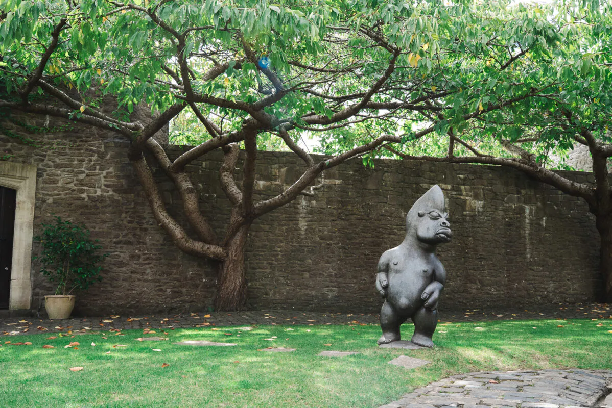 An outdoor courtyard featuring a stone wall backdrop, a large spreading tree with lush green foliage, and a distinctive stone sculpture on the grass.