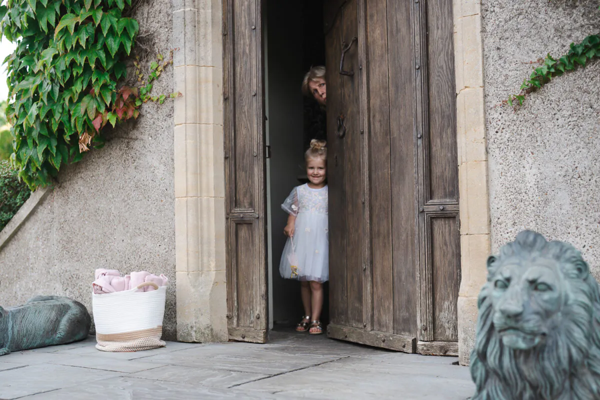 A young flower girl and a woman peek out from behind a large wooden door of a stone building with ivy on the exterior.
