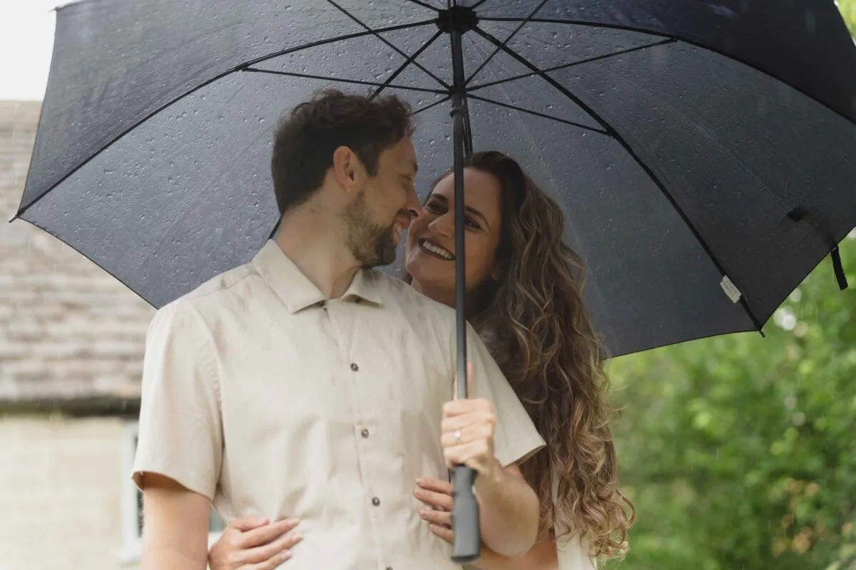 A happy couple smiling and sharing a tender moment together underneath a large black umbrella during a rainy wedding day.
