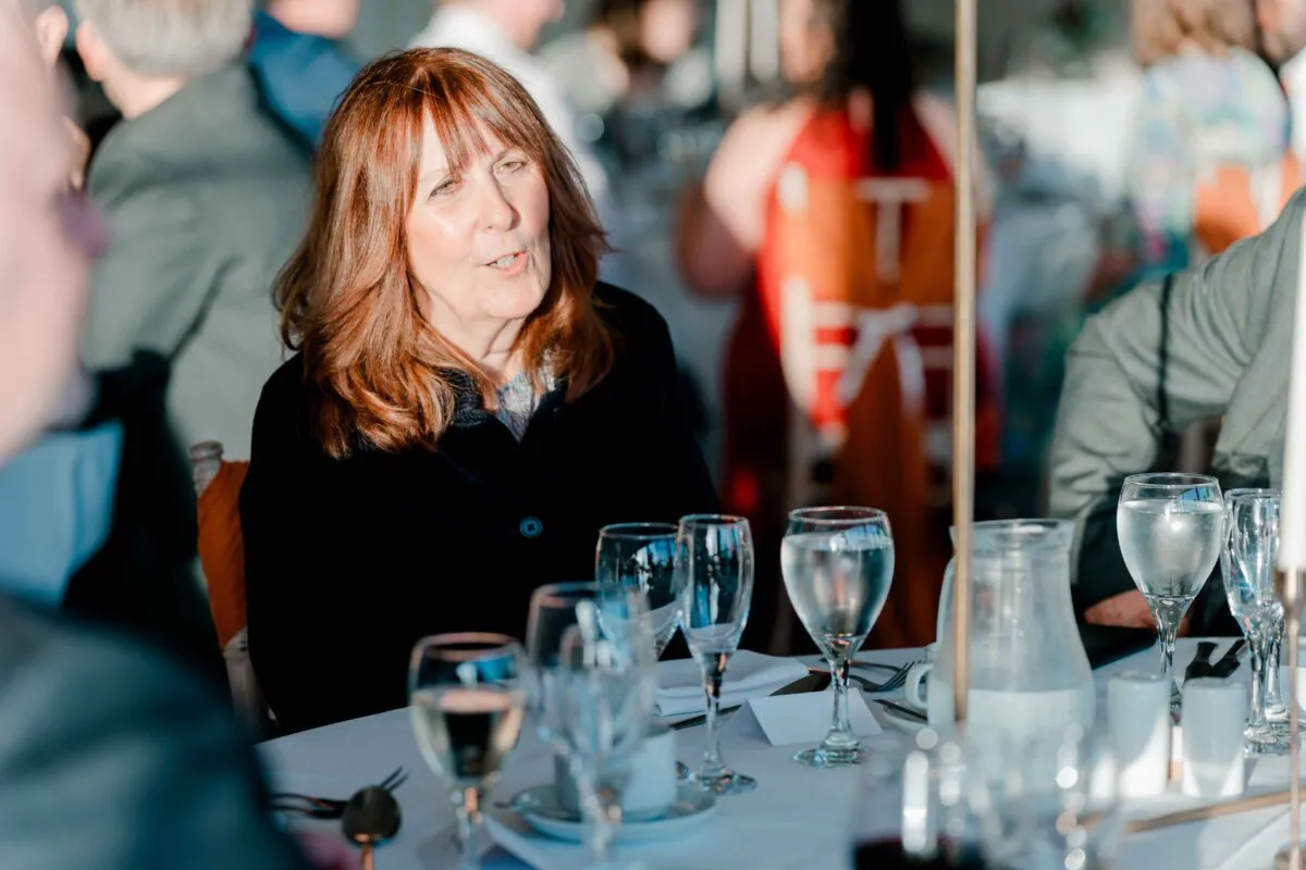 A mature female guest with reddish-brown hair is seated and engaged in conversation at a wedding breakfast reception tablescape.