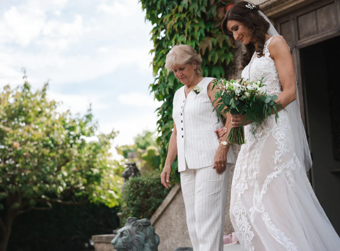 A smiling bride holding a hand-tied bouquet walks alongside her mother outdoors near a stone building with climbing ivy.