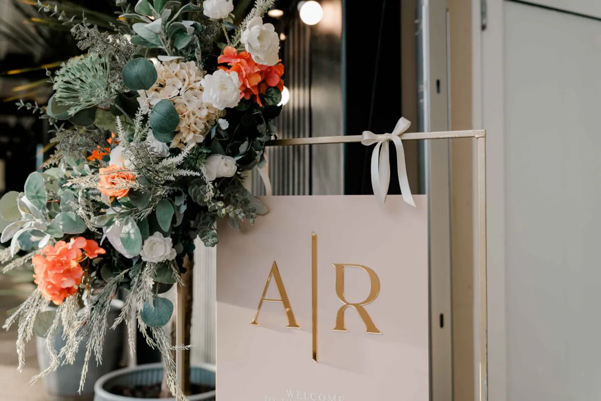 An elegant wedding welcome sign featuring gold monogram initials and a white ribbon, paired with an abundant floral arrangement containing eucalyptus and vibrant orange blooms.