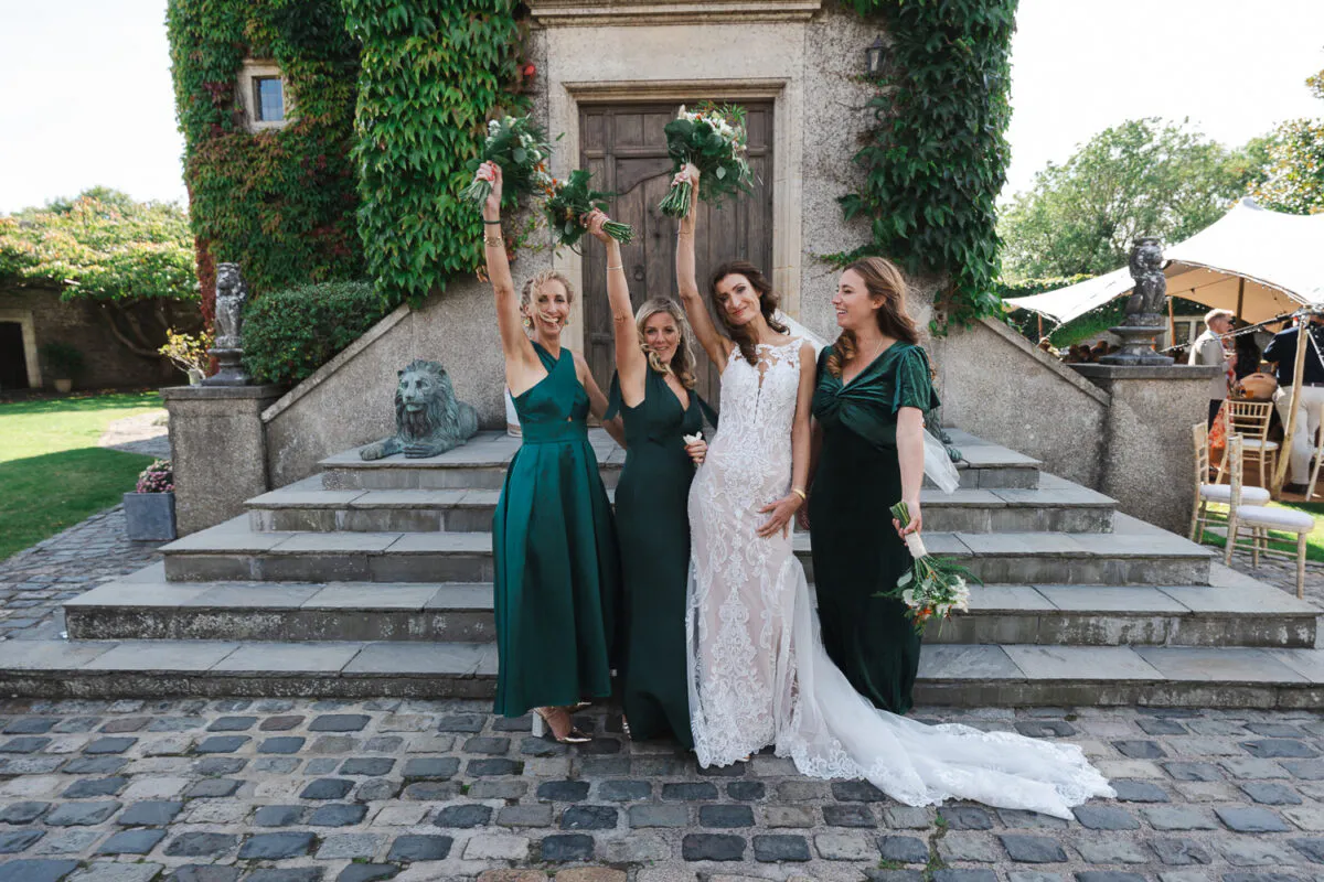 A radiant bride in a lace gown and her three bridesmaids in emerald green dresses pose joyfully on stone steps outside an ivy-clad historic venue.