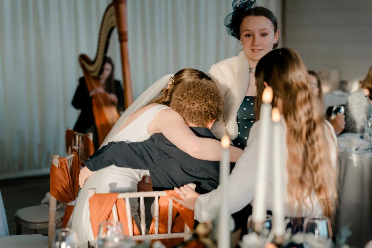 A bride shares an emotional embrace with a young child while wedding guests look on warmly in a warmly lit reception space.